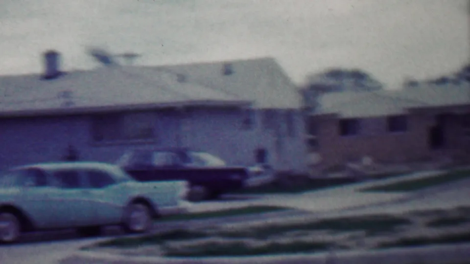 1958 Grand Rapids, Michigan home-movie frame showing parked cars along a suburban block with single-story homes, lawns, a utility pole, and soft 8mm blur.