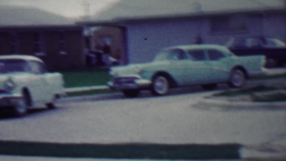 1958 Grand Rapids, Michigan frame showing two classic cars near a driveway, low residential houses, green lawns, and a paved curbside street.
