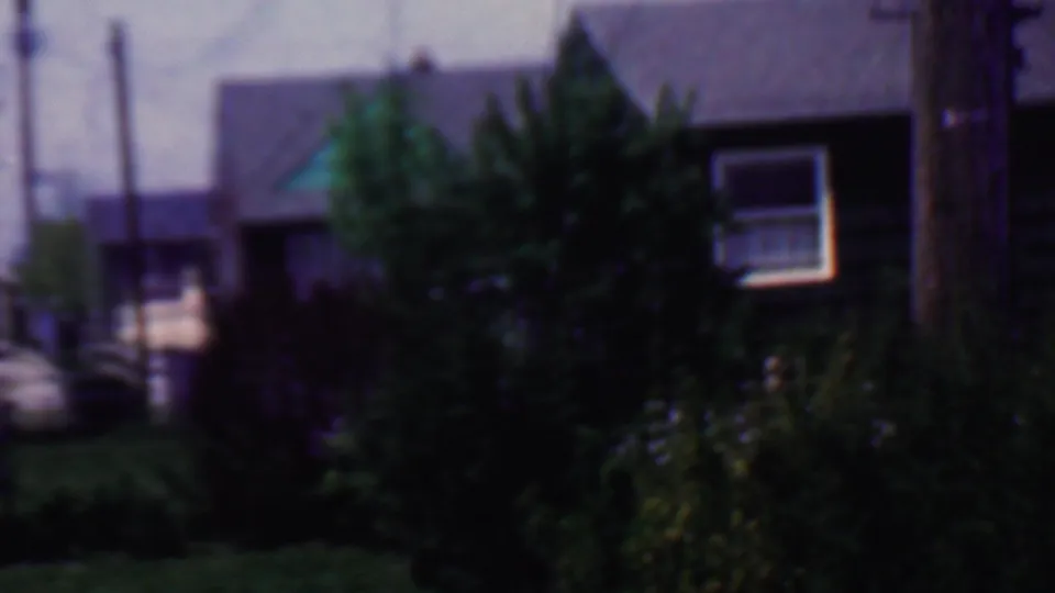 The camera shifts to a residential exterior with shrubs, a lawn, a utility pole, and a small window, consistent with the Grand Rapids home-movie setting.