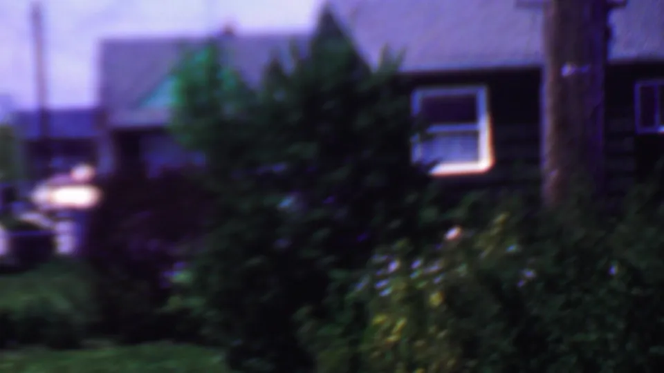 A soft-focused 8mm frame shows shrubs, a small window, lawn, and nearby residential structures in the same Grand Rapids exterior sequence.
