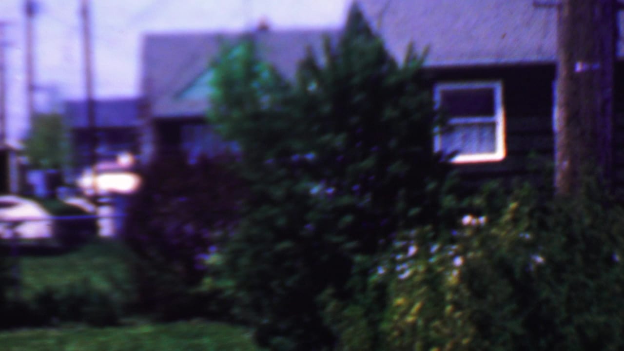 1958 Grand Rapids home-movie frame showing modest residential houses with dark roofs, green lawns, dense shrubs, utility poles, and a partly visible parked car.