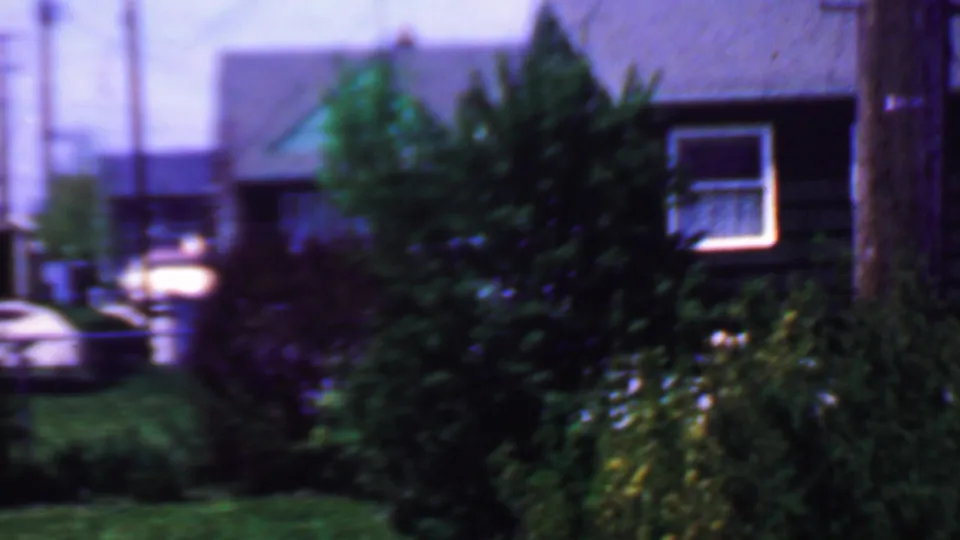 1958 Grand Rapids home-movie frame showing modest residential houses with dark roofs, green lawns, dense shrubs, utility poles, and a partly visible parked car.
