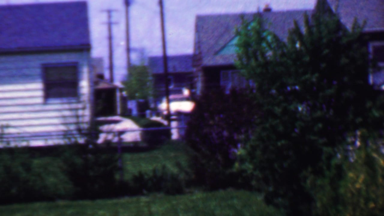 1958 Grand Rapids residential exterior with a light-sided house, dark roof, bushes, lawn, overhead utility poles, and a parked automobile near the street.