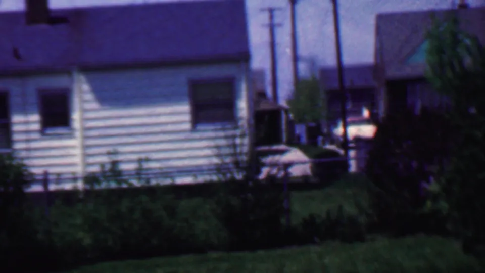 1958 Grand Rapids home-movie view of a light-colored house with horizontal siding, a dark window, shrubs, lawn, utility poles, and a parked car in the background.
