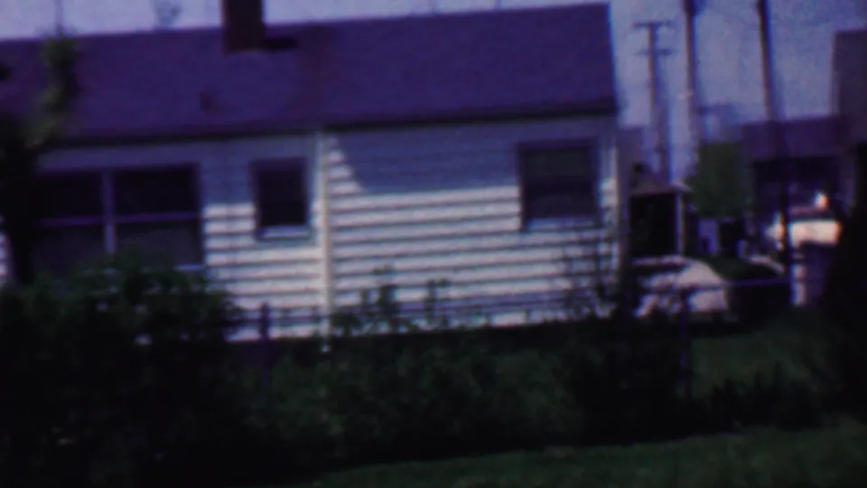 1958 Grand Rapids residential frame with a light-sided house, dark roof, windows, overgrown shrubs, lawn, utility poles, and a partial parked automobile.