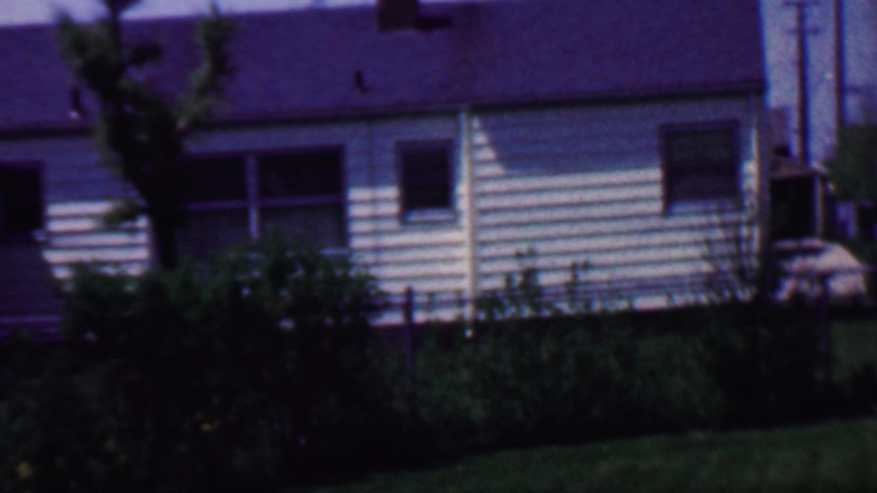 1958 Grand Rapids home-movie frame showing a white-sided suburban house with dark roof, multiple windows, chain-link fence, shrubs, and lawn in front.