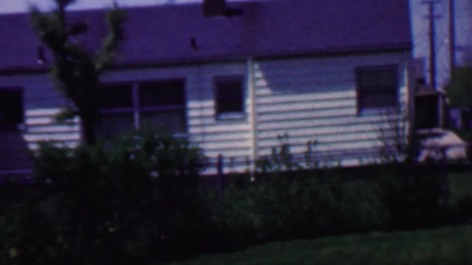 1958 Grand Rapids home-movie frame showing a white-sided suburban house with dark roof, multiple windows, chain-link fence, shrubs, and lawn in front.