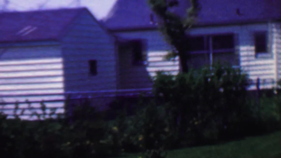 1958 Grand Rapids frame showing the camera pan moving past a white-sided house and attached shed-like section with shrubs, fencing, and lawn in the foreground.
