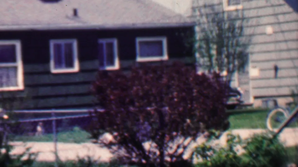 1958 Grand Rapids home-movie frame showing a darker-sided adjoining house section with windows, chain-link fence, lawn, shrubs, a small tree, and part of a bicycle at the right edge.