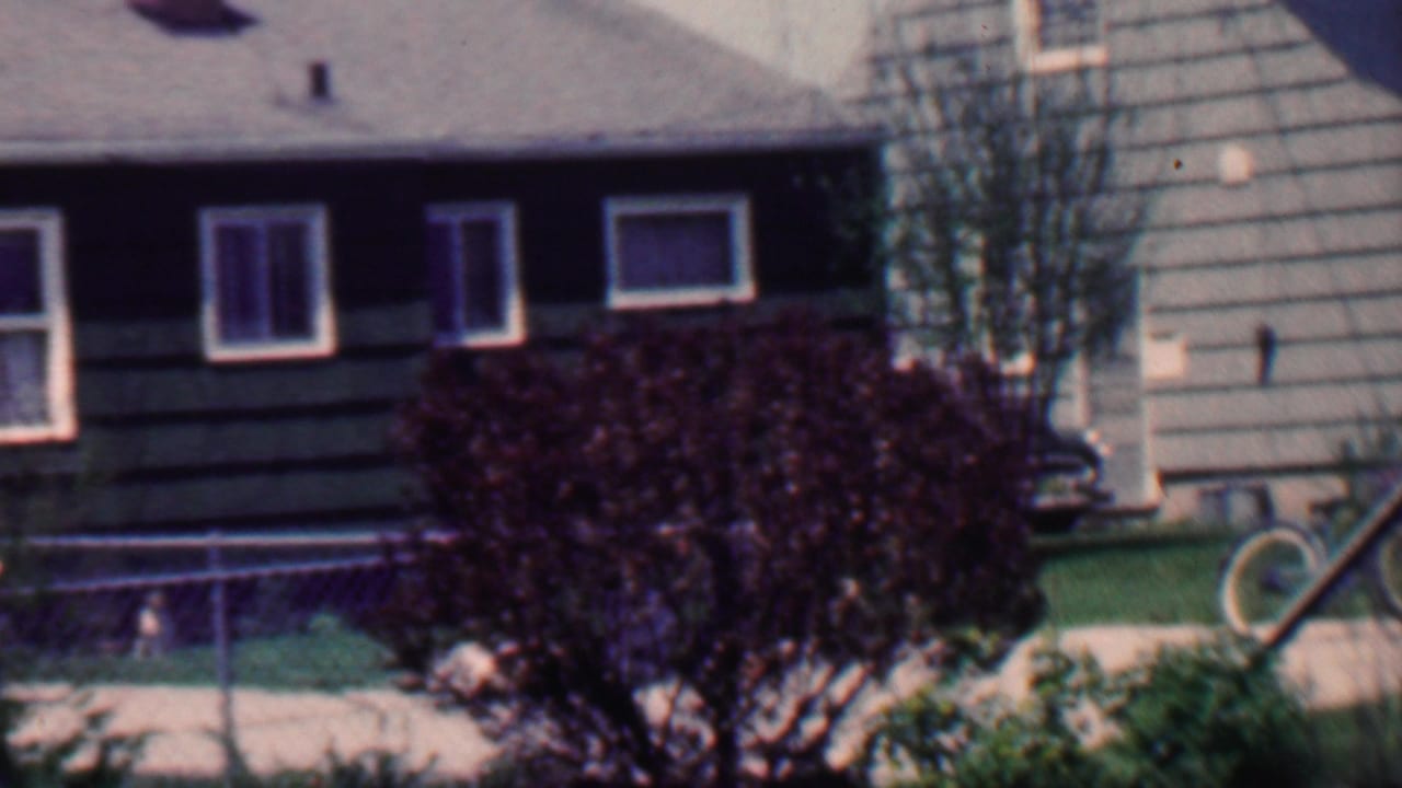 1958 Grand Rapids home-movie frame showing a suburban house with dark siding, white-trimmed windows, a chain-link fence, mowed lawn, large dark shrub, and a partial vintage car at the left edge.