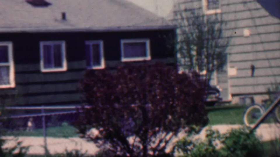 1958 Grand Rapids home-movie frame showing a suburban house with dark siding, white-trimmed windows, a chain-link fence, mowed lawn, large dark shrub, and a partial vintage car at the left edge.