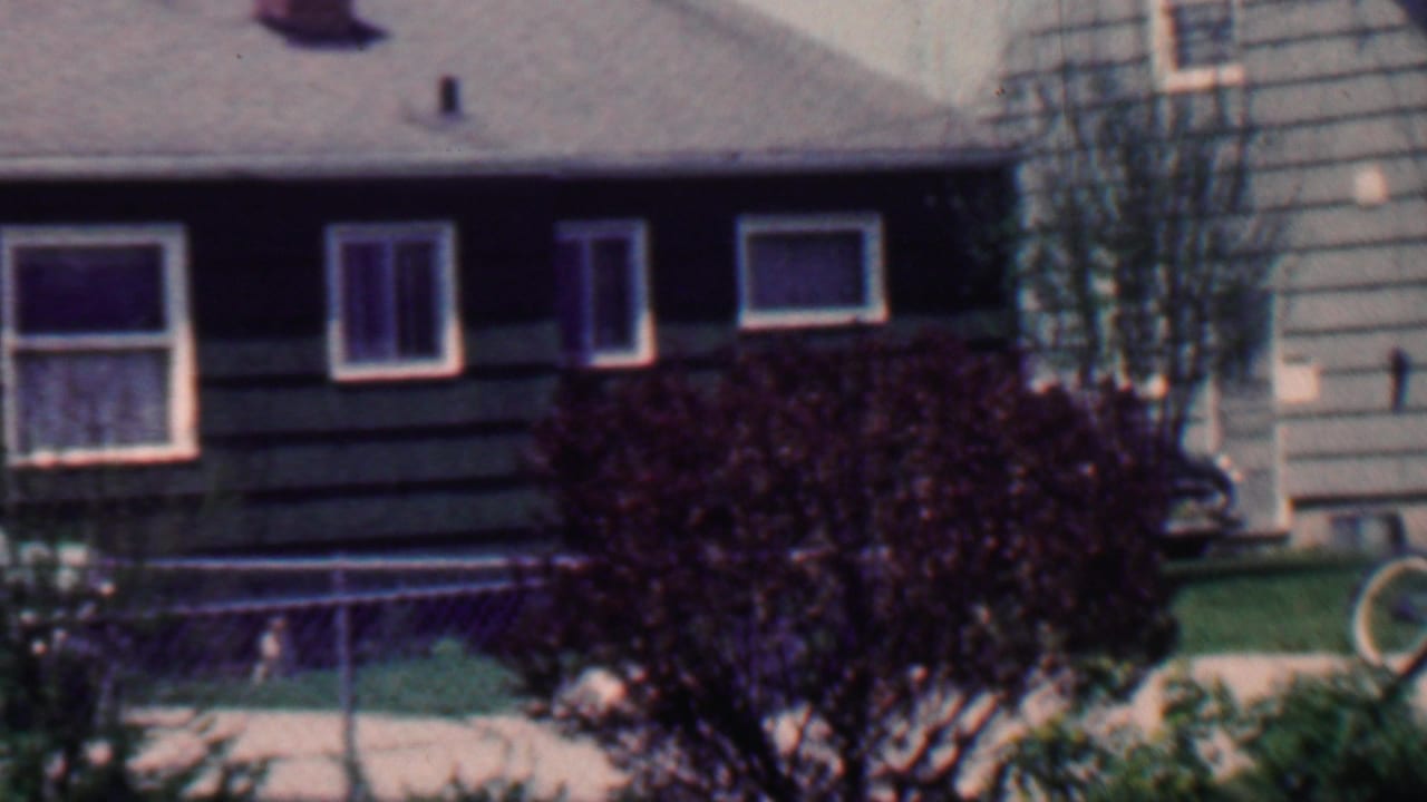 House facade with dark siding, white window trim, chain-link fence, lawn, shrub, front steps, and bicycle wheel near the right side.
