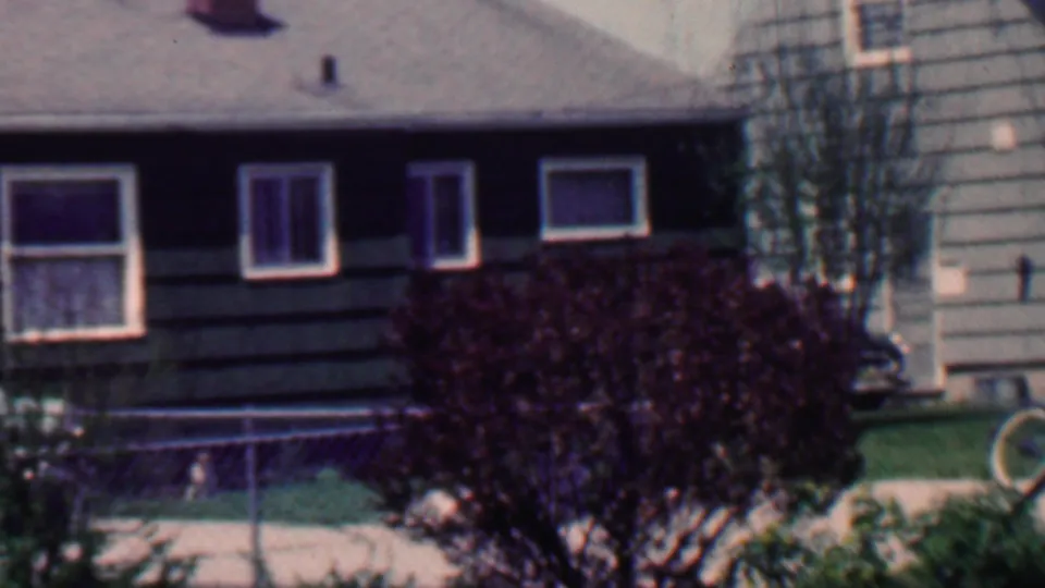 1958 Grand Rapids home-movie frame of a dark-sided suburban house with white-trimmed windows, chain-link fence, lawn, front steps, large shrub, and a bicycle wheel near the right foreground.