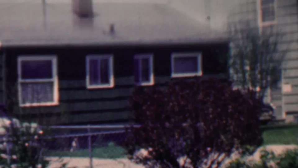 1958 Grand Rapids frame showing a slightly softer home-movie view of the suburban house front, with white-trimmed windows, chain-link fence, mowed lawn, dark shrub, and roofline visible.
