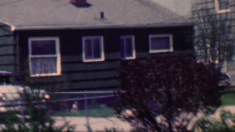 1958 Grand Rapids home-movie frame of a suburban house exterior with dark siding, white-trimmed windows, chain-link fence, front steps, mowed lawn, large dark shrub, and a thin tree at the right edge.