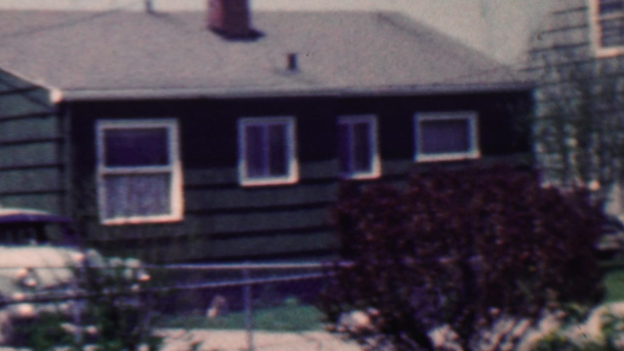 1958 Grand Rapids home-movie frame showing a dark shingled suburban house with white-trimmed windows, a chain-link fence, shrubs, lawn, and part of a car at the left edge.