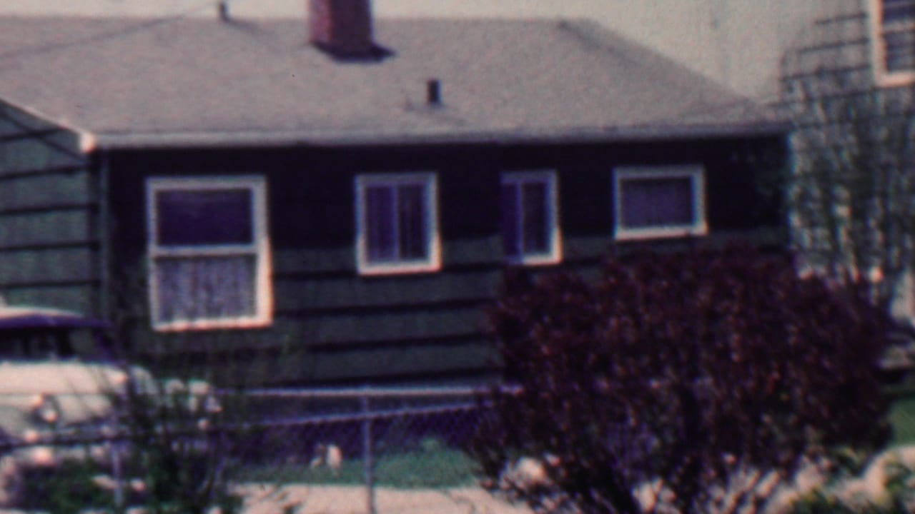1958 Grand Rapids residential exterior with a dark shingle-sided house, white window trim, chain-link fence, trimmed lawn, shrubs, and a partial parked car.