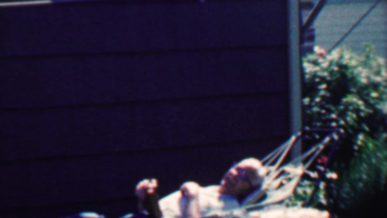 Older man with glasses lies in a hammock beside dark house siding and greenery.