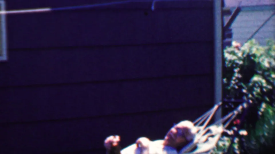 1958 Grand Rapids home-movie still showing an older man resting low in a hammock beside dark house siding and backyard greenery.