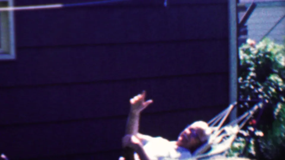 1958 Grand Rapids frame of an older man with glasses lying in a backyard hammock and raising his hand toward the home-movie camera.