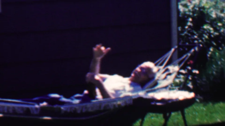 In 1958 Grand Rapids home-movie footage, an adult man reclines in a hammock beside a dark-sided house and raises one hand in a casual wave.