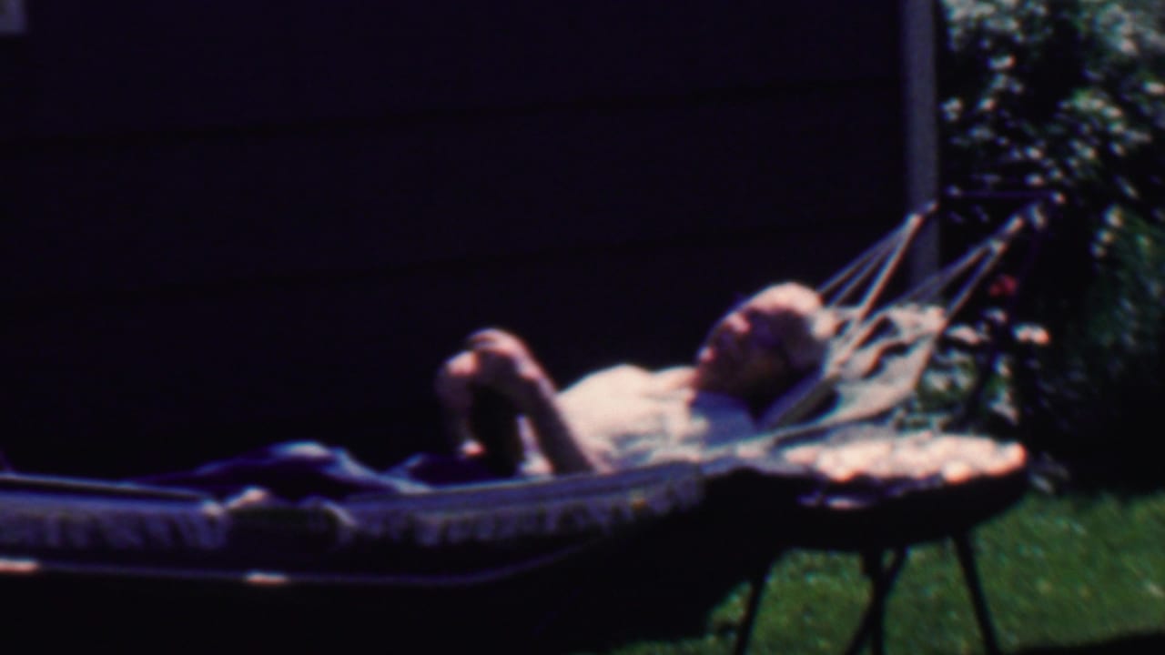 The man remains reclined in the backyard hammock, with dark house siding, lawn, and shrubs visible in the 1958 Grand Rapids home-movie frame.