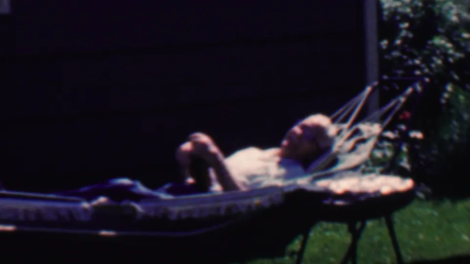 The man remains reclined in the backyard hammock, with dark house siding, lawn, and shrubs visible in the 1958 Grand Rapids home-movie frame.