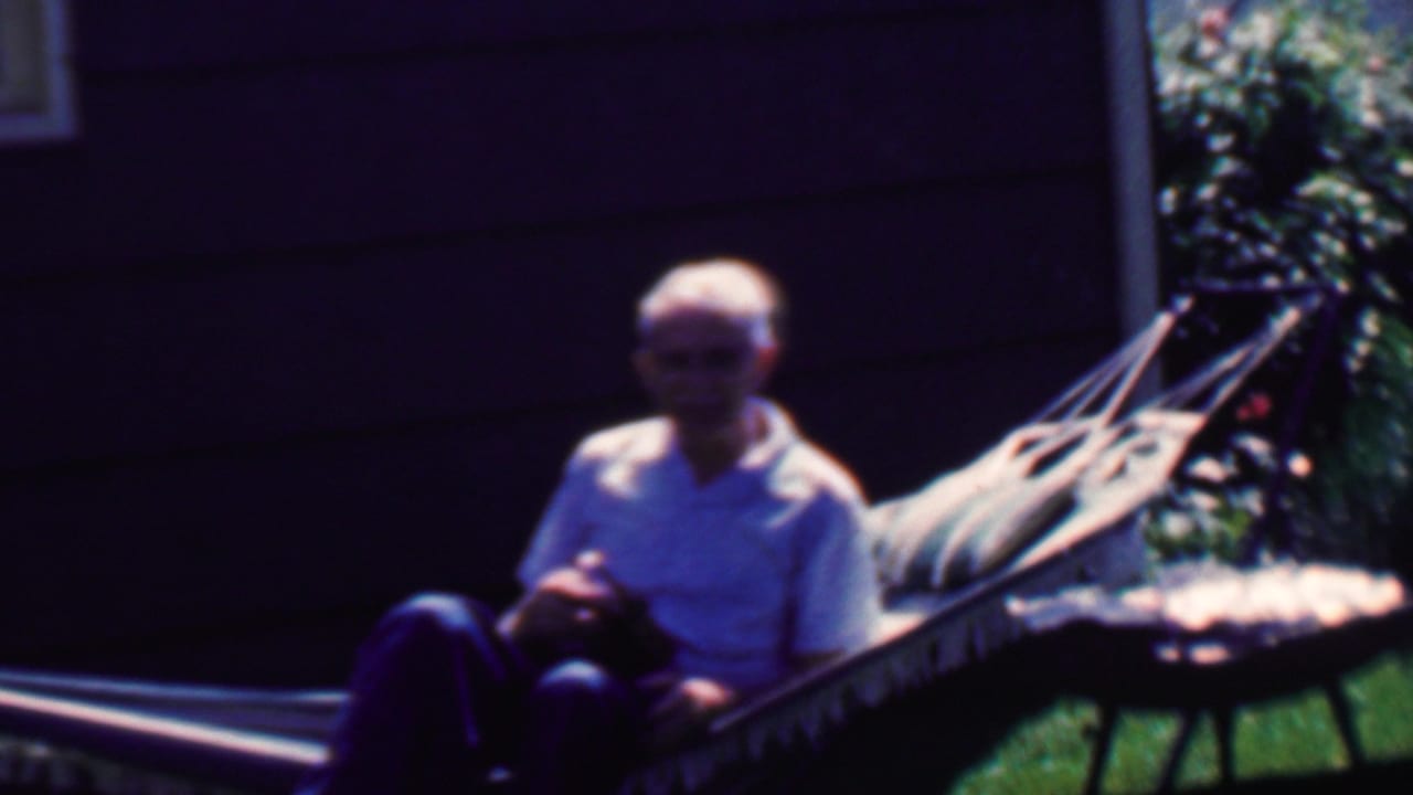 In 1958 Grand Rapids home-movie footage, an elderly man sits in a fabric hammock on a sunny lawn near dark house siding and a small round grill or table.
