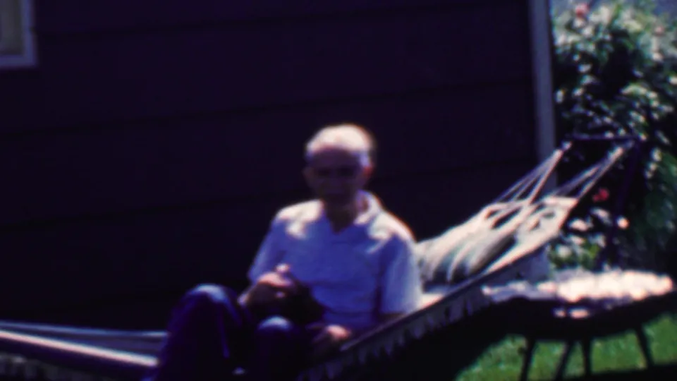 In 1958 Grand Rapids home-movie footage, an elderly man sits in a fabric hammock on a sunny lawn near dark house siding and a small round grill or table.
