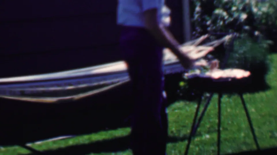 An elderly man stands beside a hammock and small round grill or table in a sunlit Grand Rapids backyard, with lawn and shrubs visible behind him.