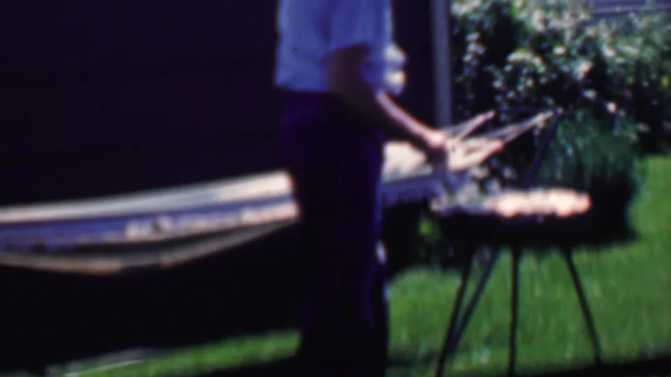 The man stands close to the small round grill or table with the hammock stretched behind him, captured in soft 8mm color in a sunny residential yard.