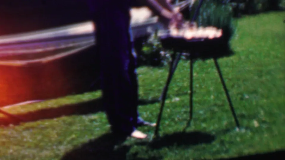 A cropped view shows the man beside the small round grill or table, with the hammock, green lawn, and a warm film flare at the edge of the frame.
