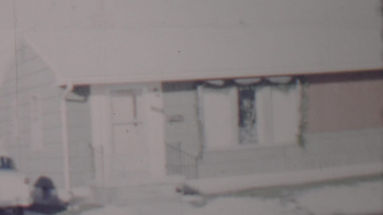 Soft 8mm frame of a snow-covered suburban house in Lincoln, Nebraska, according to metadata, with a front door, curtained window, and a car cropped at the driveway edge.
