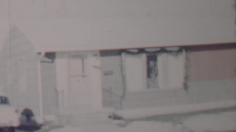 Soft 8mm frame of a snow-covered suburban house in Lincoln, Nebraska, according to metadata, with a front door, curtained window, and a car cropped at the driveway edge.