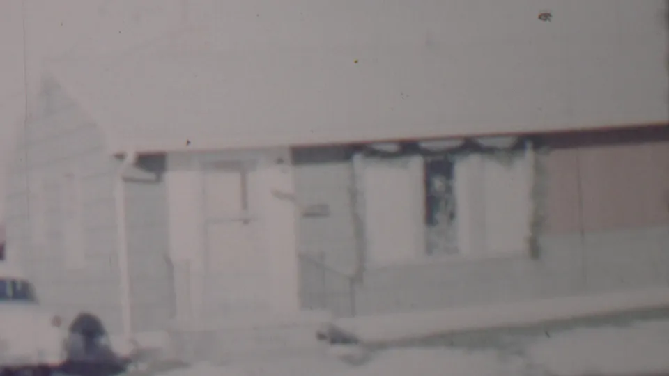 Soft-focus 8mm still of the snow-covered residential facade, with the driveway car partly cropped, the front entry, and window details visible in the frame.