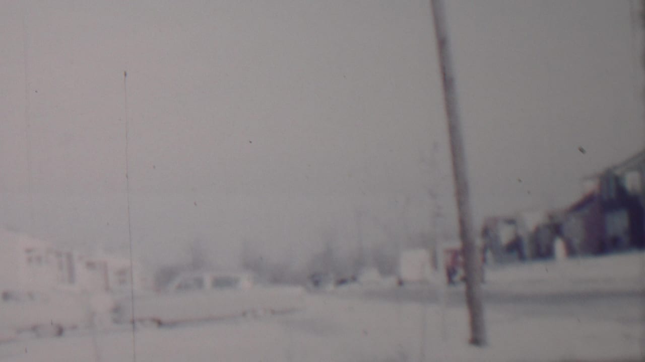 Soft 8mm view of a snow-covered residential street in Lincoln, Nebraska, with parked cars, modest houses, bare trees, and a foreground telephone pole.
