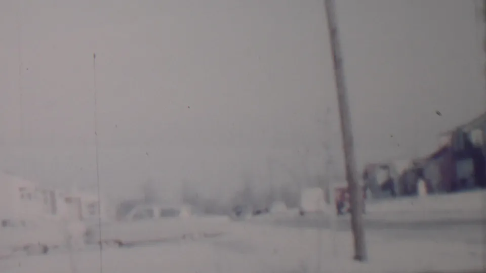 Soft 8mm view of a snow-covered residential street in Lincoln, Nebraska, with parked cars, modest houses, bare trees, and a foreground telephone pole.
