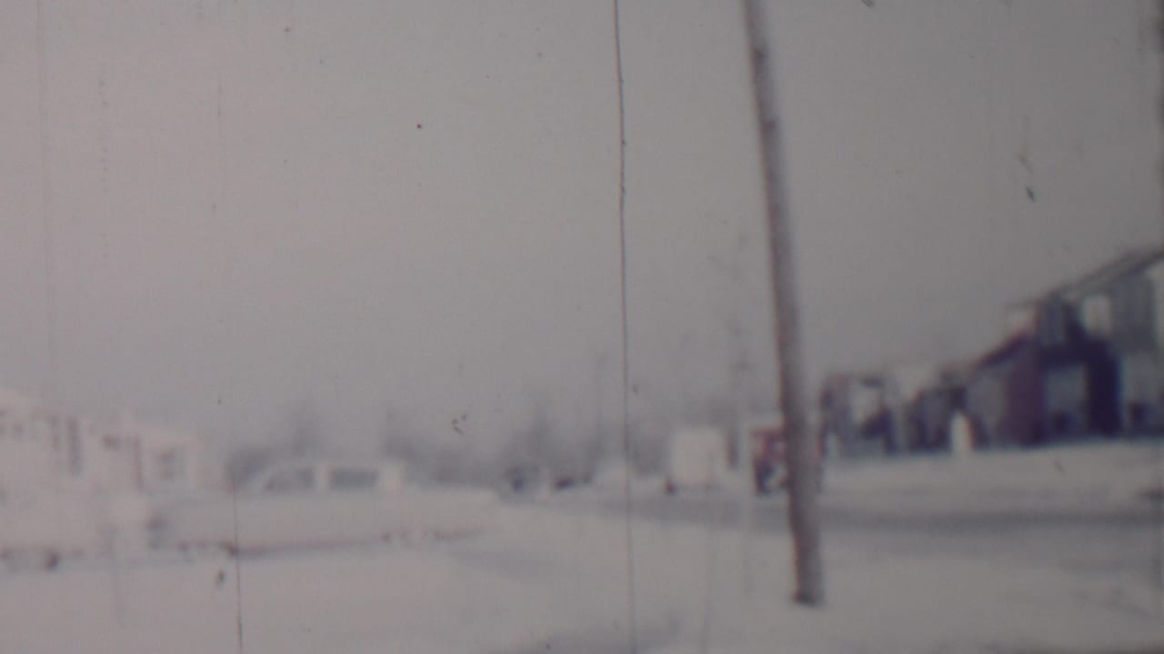 Snowy street view with houses, parked cars, and a utility pole near the foreground.