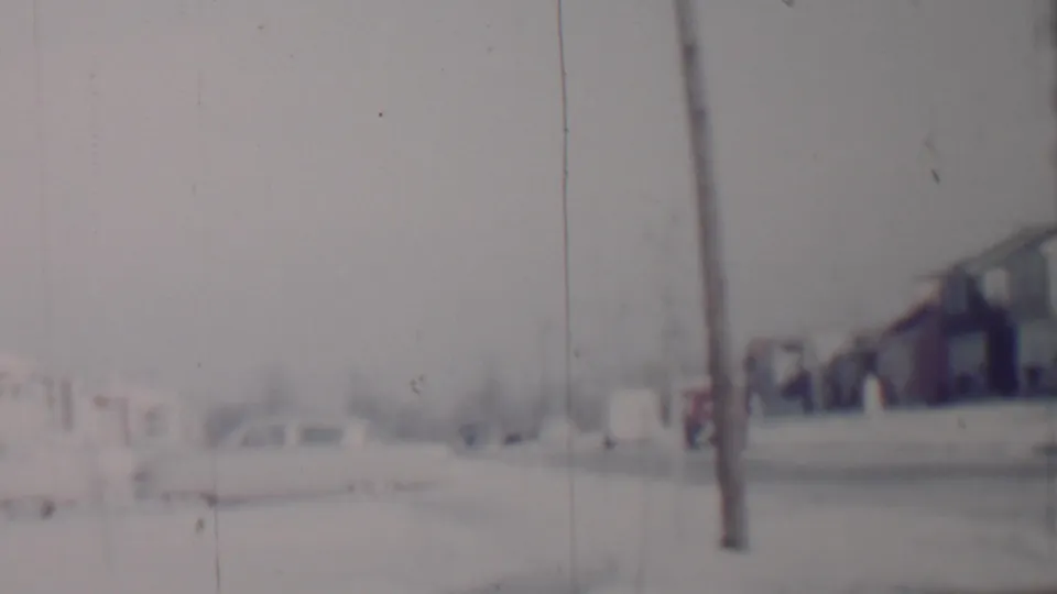 Overexposed home-movie frame of a Lincoln, Nebraska winter neighborhood, showing a utility pole, snowy pavement, parked cars, and detached houses.