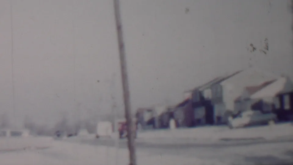 Grainy 8mm frame of a snow-covered Lincoln residential block, with a foreground pole, parked cars, distant houses, and winter trees.