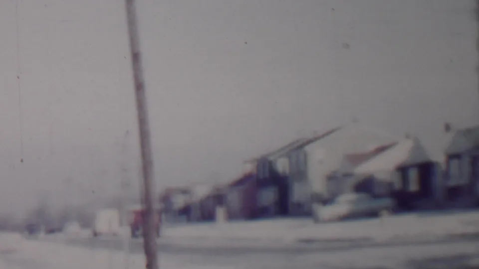 Soft, grainy Lincoln, Nebraska home-movie frame showing a snowy suburban street, telephone pole, parked cars, and low residential houses.