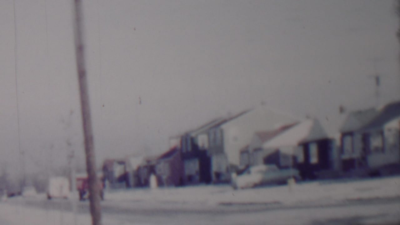 Soft 8mm frame of a 1962 Lincoln, Nebraska suburban street with snow on lawns and rooftops, modest houses, parked cars, and a foreground utility pole.
