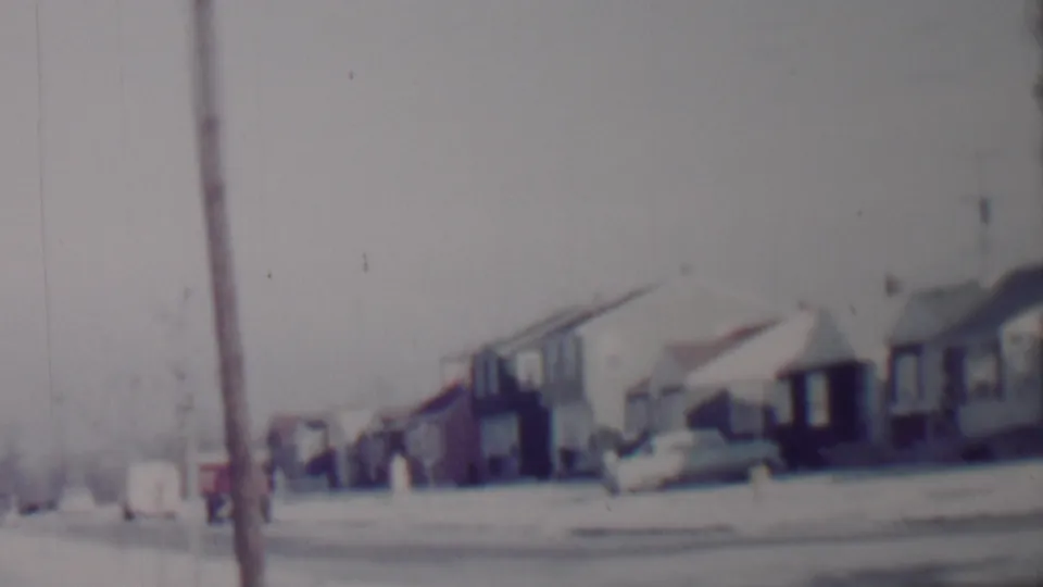 Soft 8mm frame of a 1962 Lincoln, Nebraska suburban street with snow on lawns and rooftops, modest houses, parked cars, and a foreground utility pole.