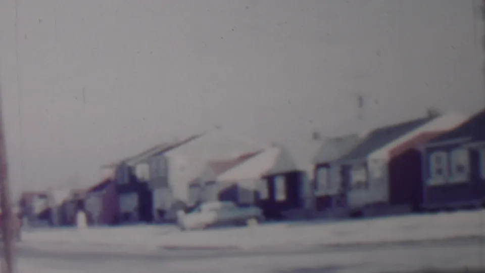 Faded-color 8mm view of a snow-covered residential block in Lincoln, Nebraska, with low houses, parked mid-century cars, and utility lines.