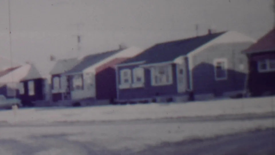 Final 8mm frame of the pan across a 1962 Lincoln, Nebraska winter street, showing modest homes, snow on roofs and lawns, and a parked car near the curb.
