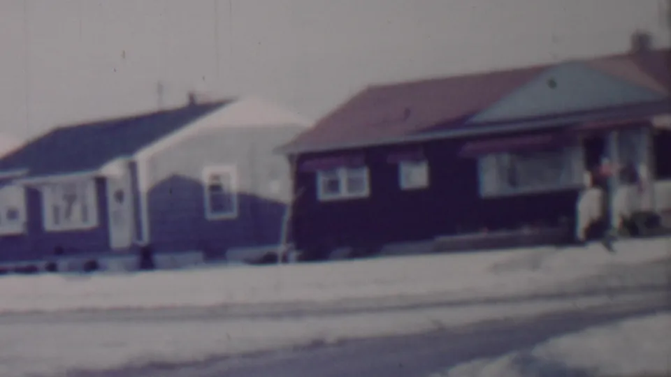 A snowy Lincoln, Nebraska residential street appears in soft 8mm footage, with bungalows in view and a person briefly visible near a porch on the right.