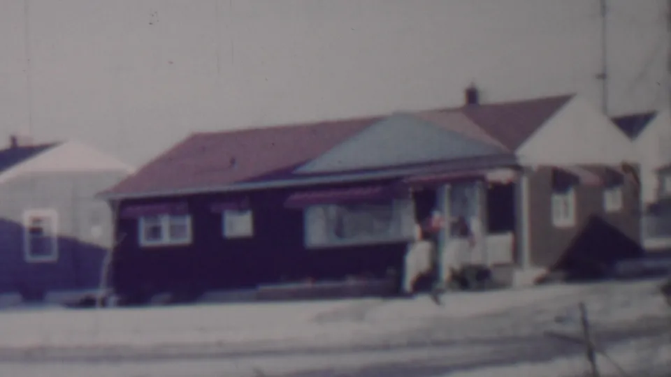 A late frame of the 1962 Lincoln, Nebraska home-movie pan shows snow along the curb, bungalow facades, and a person near a porch at the right side of the scene.