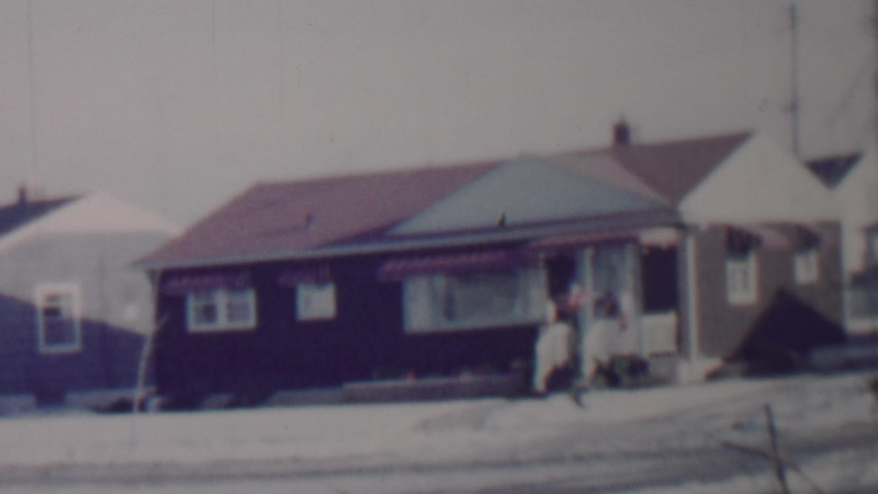 1962 Lincoln, Nebraska home-movie frame showing a modest single-story house with dark siding, a red roof, snow on the ground, and a person standing near the door.