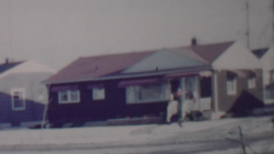 1962 Lincoln, Nebraska home-movie frame showing a modest single-story house with dark siding, a red roof, snow on the ground, and a person standing near the door.