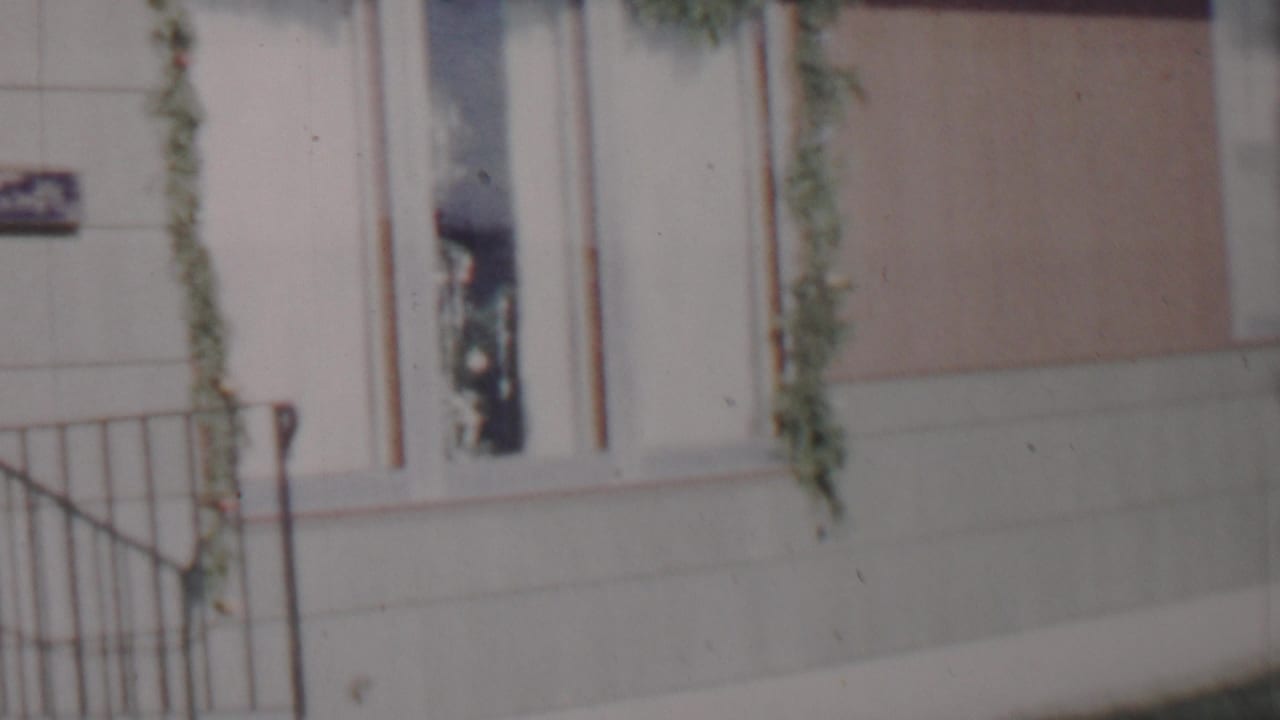 Close 1962 Lincoln, Nebraska frame of a house entry area with metal porch railings, a light-colored door, a narrow window, green garland, and a partially visible sign.